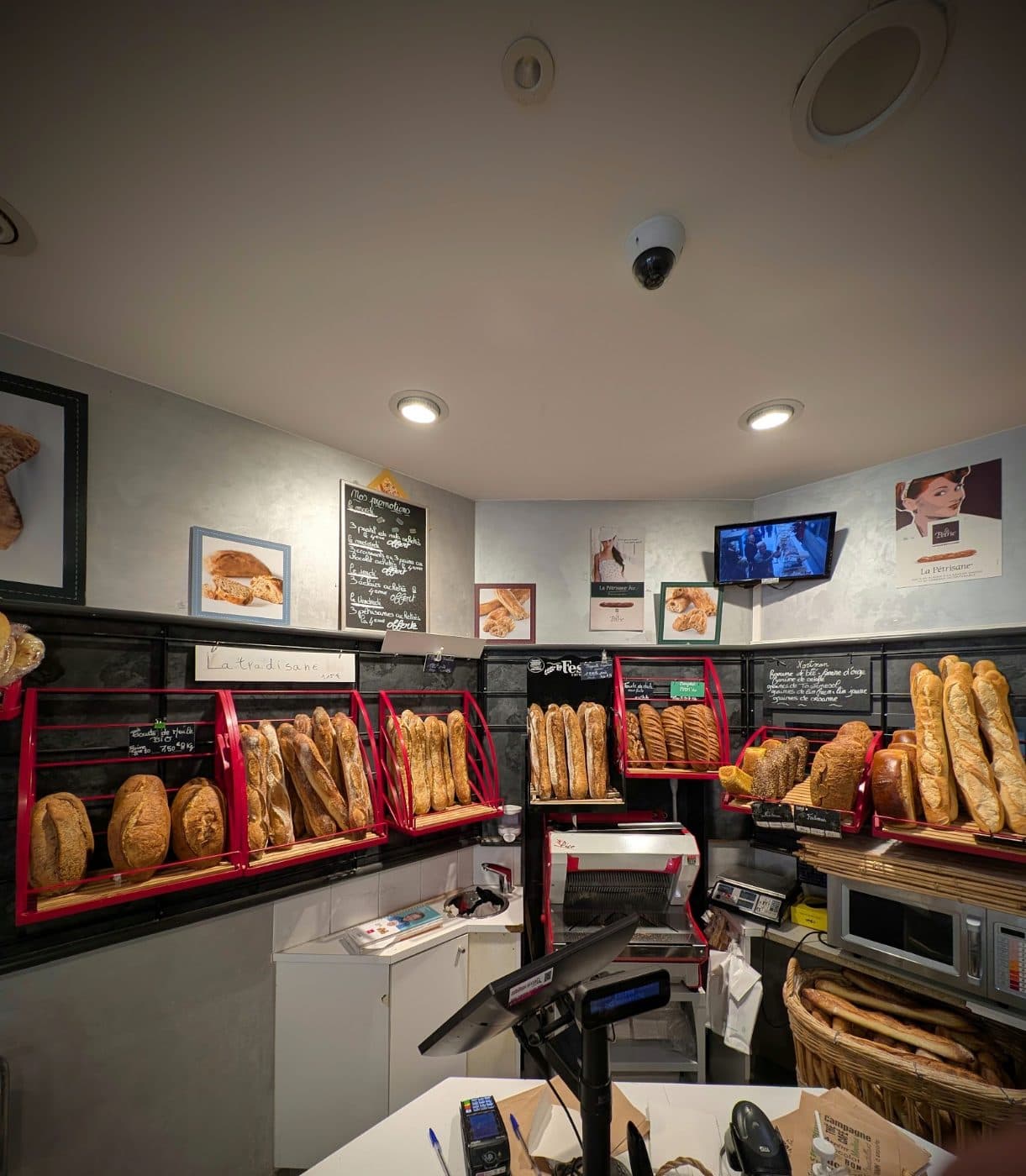 L’intérieur de la boutique : casiers rouges garnis de baguettes et de pains le long du mur, trancheuse au fond.